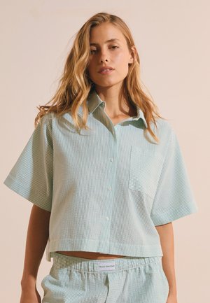 Giovane donna con capelli biondi sciolti che indossa una camicia a maniche corte a righe bianche e verde chiaro con bottoni e pantaloni abbinati.
