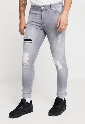 Mand iført lysegrå, slidte skinny jeans, hvide strømper og sorte sneakers, stående foran en ensfarvet baggrund.
