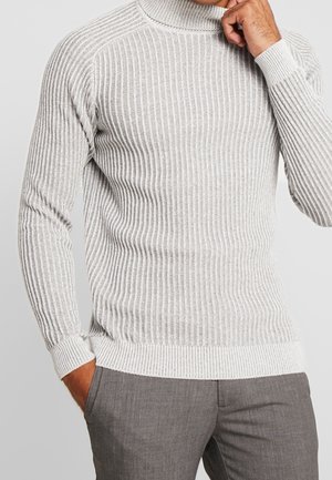 Homme portant un pull à col roulé côtelé gris clair et un pantalon gris, une main dans la poche et l'autre près du menton.