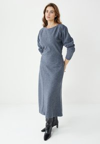 Robe longue ajustée en tissu gris texturé. Elle présente un col rond, des manches longues et des poches latérales. Associée à des bottines noires.