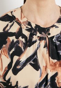 Blouse beige avec motif abstrait de coups de pinceau noirs, marron et gris, col rond et deux boutons noirs près de l'encolure.