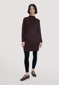 Strickkleid in dunklem Burgunderrot mit einem hohen Kragen, langen Ärmeln und seitlichen Schlitzen, kombiniert mit schwarzen Leggings und braunen Ballerinas.