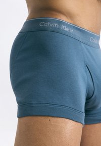 Vīrietis, kurš valkā zilas Calvin Klein bokseršortus, parāda jostasvietu un sānu profilu uz vienkārša gaiša fona.