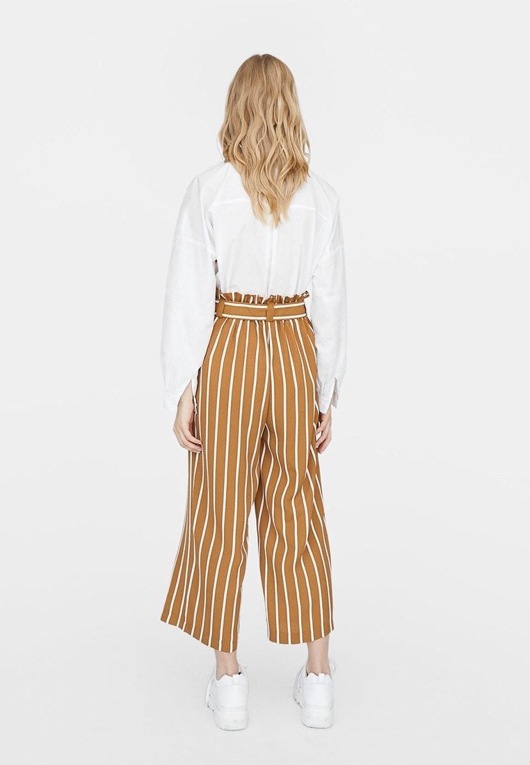 Pantalon à jambes larges rayé marron et blanc avec une taille ceinturée, associé à un haut blanc ample et des chaussures blanches.