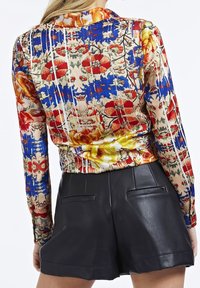 Blouse à manches longues avec un motif floral et abstrait dans des couleurs vives ; dotée d'un col et d'un nœud à la taille, associée à un short en cuir noir.