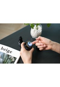 Svart glasskrukke med blå krem inni, hånd som påfører kremen, svart pipettflaske i nærheten, åpen magasin med bilde av plante og "beige" tekst.
