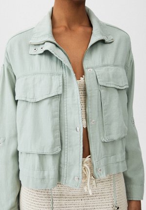 Woman wearing a light green cropped jacket with large front pockets over a beige knit top and matching knit pants.