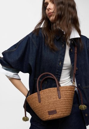 Mujer con chaqueta vaquera oscura y camisa blanca, sosteniendo un bolso marrón tejido con asas de cuero y correa ajustable.