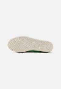 Green sneaker with a textured white rubber sole, featuring circular grips and embossed brand name. Smooth and durable upper material.