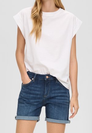 Femme portant un short en denim bleu foncé retroussé et un t-shirt blanc sans manches, debout avec une main dans la poche.