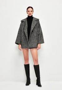 Cappotto oversize a scacchi grigi con ampi risvolti, tessuto testurizzato, chiusura con un solo bottone, indossato su un dolcevita nero e un completo corto.