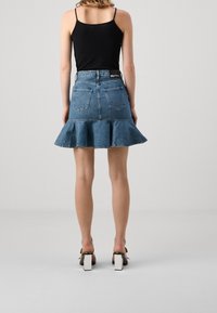 Vrouw in een zwart mouwloos topje, met een blauw spijkerrok met ruches en achterzakken, en zwarte sandalen met witte blokhakken, staand met haar rug naar de kijker gekeerd.