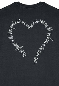 Melna T-krekls ar baltu tekstu, kas veido sirds formu uz muguras, ar roku rakstītu ziņu par vienotību dabā.