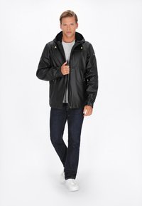 Giovane uomo che indossa una giacca nera con cappuccio, una maglietta grigia, jeans scuri e sneaker bianche, in piedi mentre sistema la cerniera della giacca.