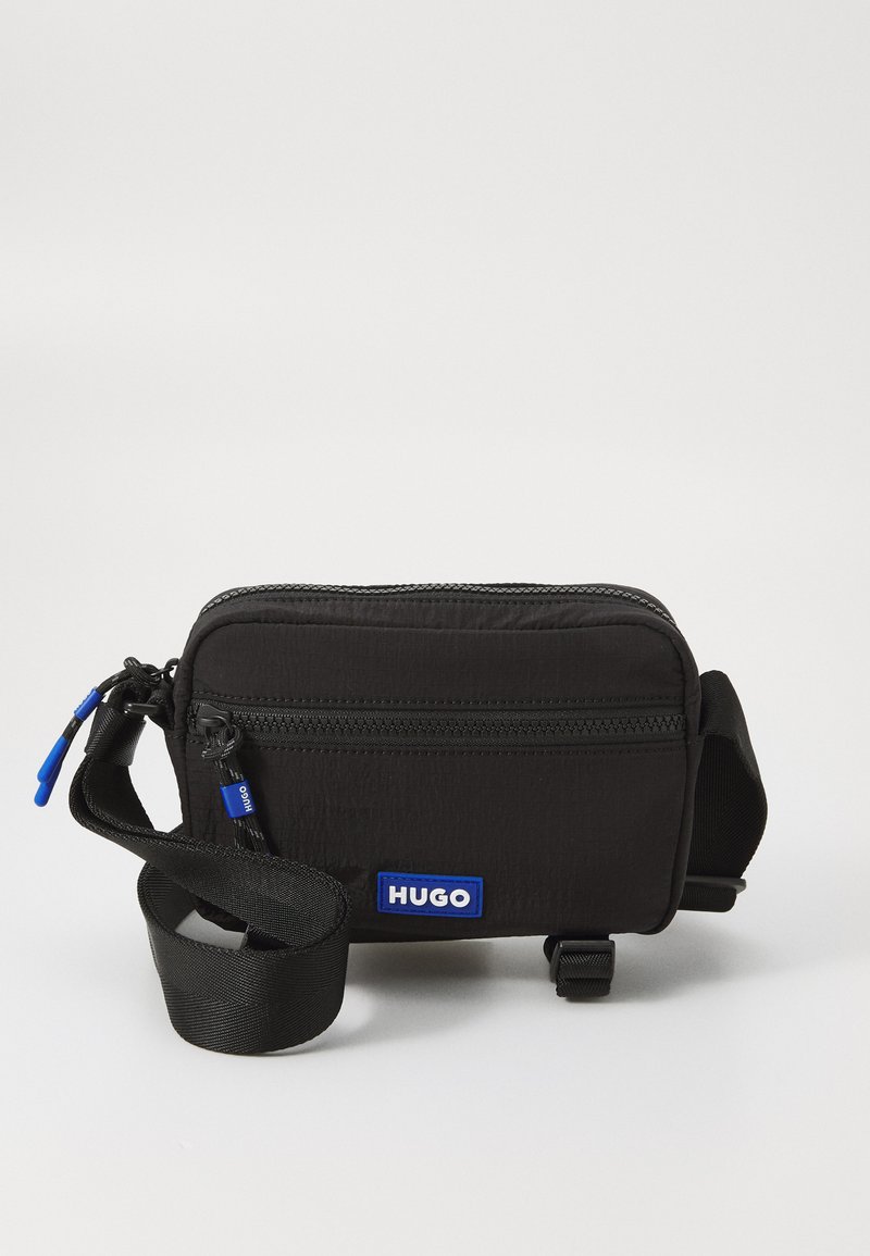 Borsa a tracolla in tessuto nero con chiusura a zip, toppa con logo blu, tracolla regolabile e superficie testurizzata. Design compatto e rettangolare.