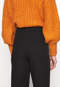 Oranje gebreide sweater met oversized mouwen, gecombineerd met zwarte op maat gemaakte broeken. Gelaagde stof en strakke lijnen op de broek.