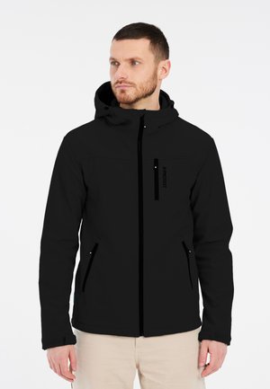 Mężczyzna o krótkich brązowych włosach i brodzie, ubrany w czarną kurtkę z kapturem z kieszeniami na zamek błyskawiczny oraz beżowe spodnie, patrzący w lewo na białym tle.