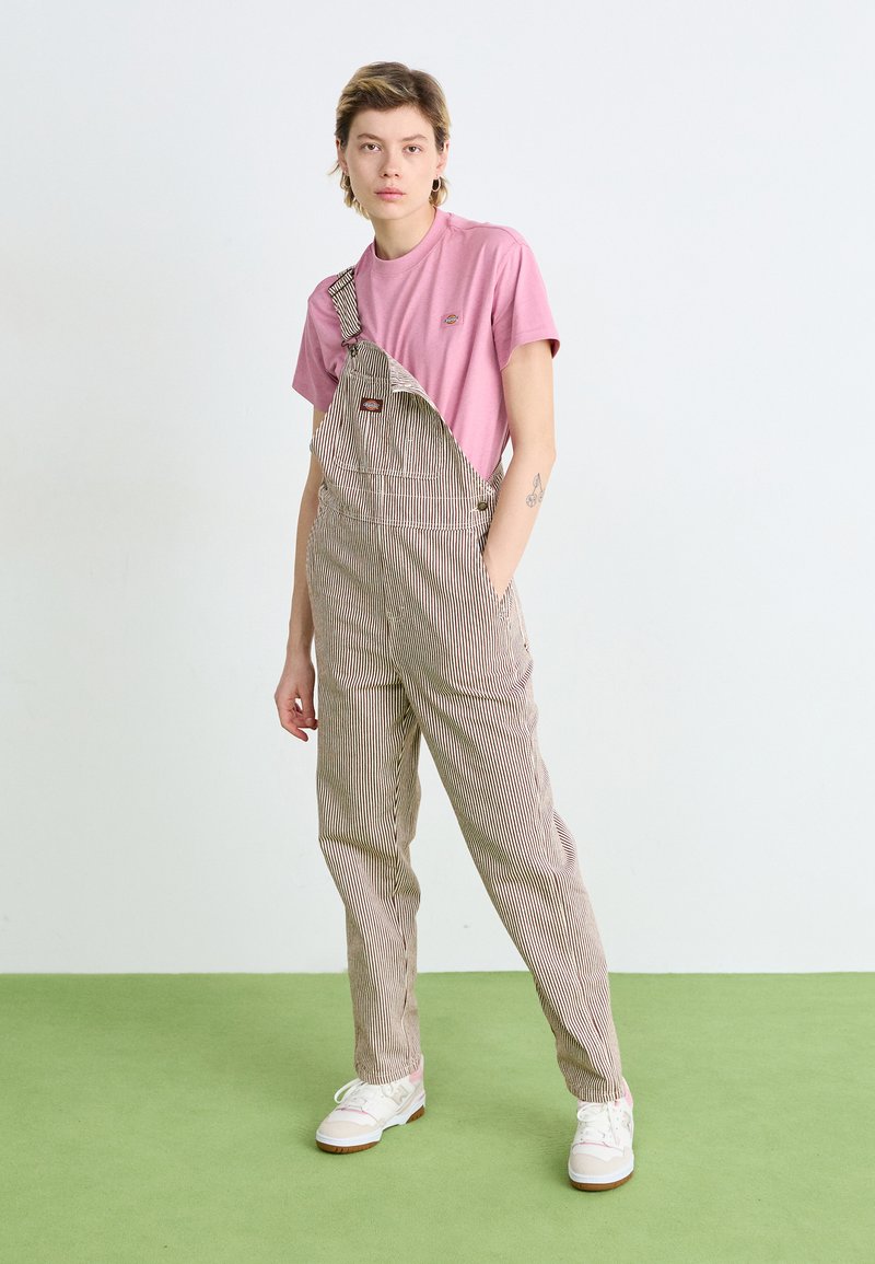 Salopette a righe beige con tasca, indossata sopra una maglietta rosa. Il modello si trova su un pavimento verde, indossando sneaker bianche.