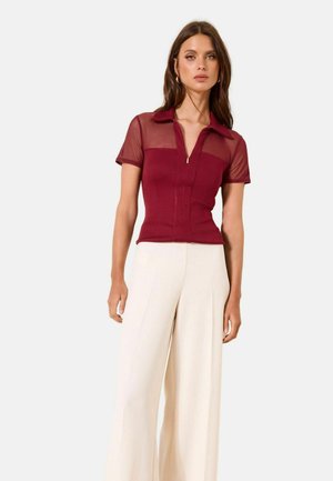 Žena má na sobě přiléhavé vínové tričko s krátkými rukávy, průhlednými panely a límečkem, doplněné o krémové široké kalhoty s vysokým pasem.