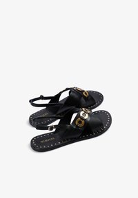 Sandali neri piatti con cinturini incrociati, dettagli in metallo dorato e applicazioni di borchie sulla suola. Realizzati in pelle liscia.
