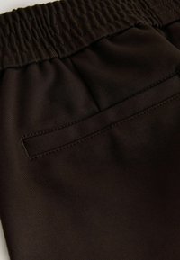 Close-up of dark brown elastic waistband and back welt pocket on smooth fabric trousers.
