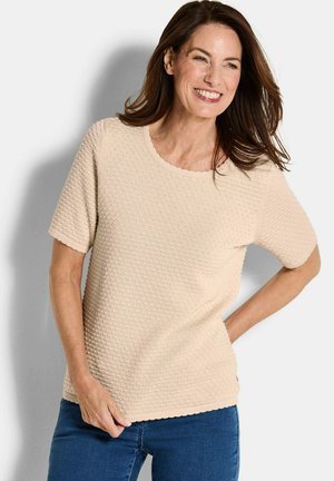 Frau lächelt, trägt ein beigefarbenes, strukturiertes Kurzarmoberteil und blaue Jeans, steht mit einer Hand an der Taille vor einem weißen Hintergrund.