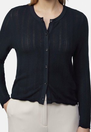 Femme portant un cardigan texturé noir à manches longues boutonné et un pantalon beige clair, mains dans les poches, torse visible.