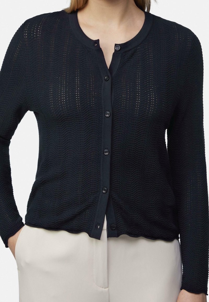 Femme portant un cardigan texturé noir à manches longues boutonné et un pantalon beige clair, mains dans les poches, torse visible.