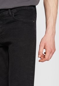 Juodi džinsai su siauru kirpimu, turintys priekinės kišenes ir subtilias siuvimo detales. Ranka remiasi į šlaunies sritį.