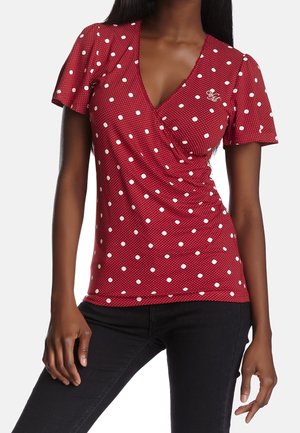 Kobieta nosząca czerwoną bluzkę z krótkim rękawem i dekoltem w serek w białe kropki oraz czarne dopasowane spodnie, stojąca z jedną ręką opuszczoną wzdłuż ciała.