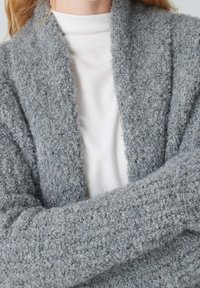 Persona vestida con un suéter de cuello alto blanco y un cárdigan de lana gris texturizado, con los brazos cruzados, cortado a la altura de los hombros y el cuello.
