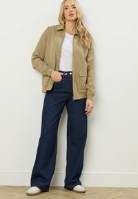 Giacca khaki con zip e colletto, abbinata a jeans in denim scuro a gamba larga e scarpe bianche slip-on. Tessuto liscio e design strutturato.