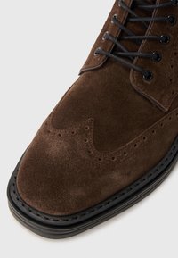 Botín de ante marrón hasta el tobillo con detalles de brogue, ojales negros y cordones negros. Presenta una puntera redonda y suela de goma negra en contraste.