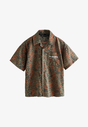 Jugendlicher Junge mit kurzen lockigen Haaren trägt ein orange-grünes, kurzärmeliges Hemd mit Leopardenmuster über einem schwarzen T-Shirt und steht vor einem grauen Hintergrund.