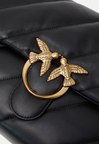 Zwarte gestikte leren handtas met een circulaire gouden sluiting met twee gestileerde vogels, wat een getextureerd ontwerp element toevoegt.