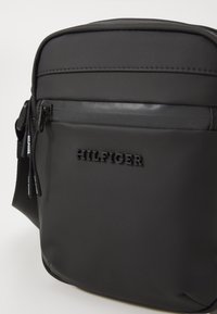 Geantă de umăr din nailon negru, cu logo-ul „HILFIGER” embosat, textură netedă, curea ajustabilă și un buzunar frontal cu fermoar.