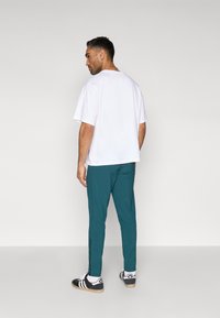 Fehér rövid ujjú póló, türkizszínű slim-fit nadrág, és fekete, fehér részletekkel ellátott sportcipő. Látható cipzár részlet a nadrágvarrás mentén.