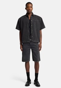Camicia a maniche corte in denim nero con taschino sul petto, abbinata a pantaloni shorts in denim nero. Indossata con una canotta nera e mocassini neri.