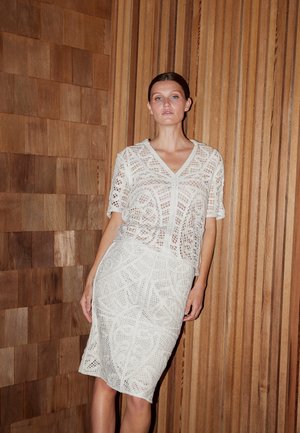 Femeie cu păr închis la culoare poartă top alb de croșetat asortat cu fustă, stând în fața unui perete din lemn cu panouri verticale și orizontale.