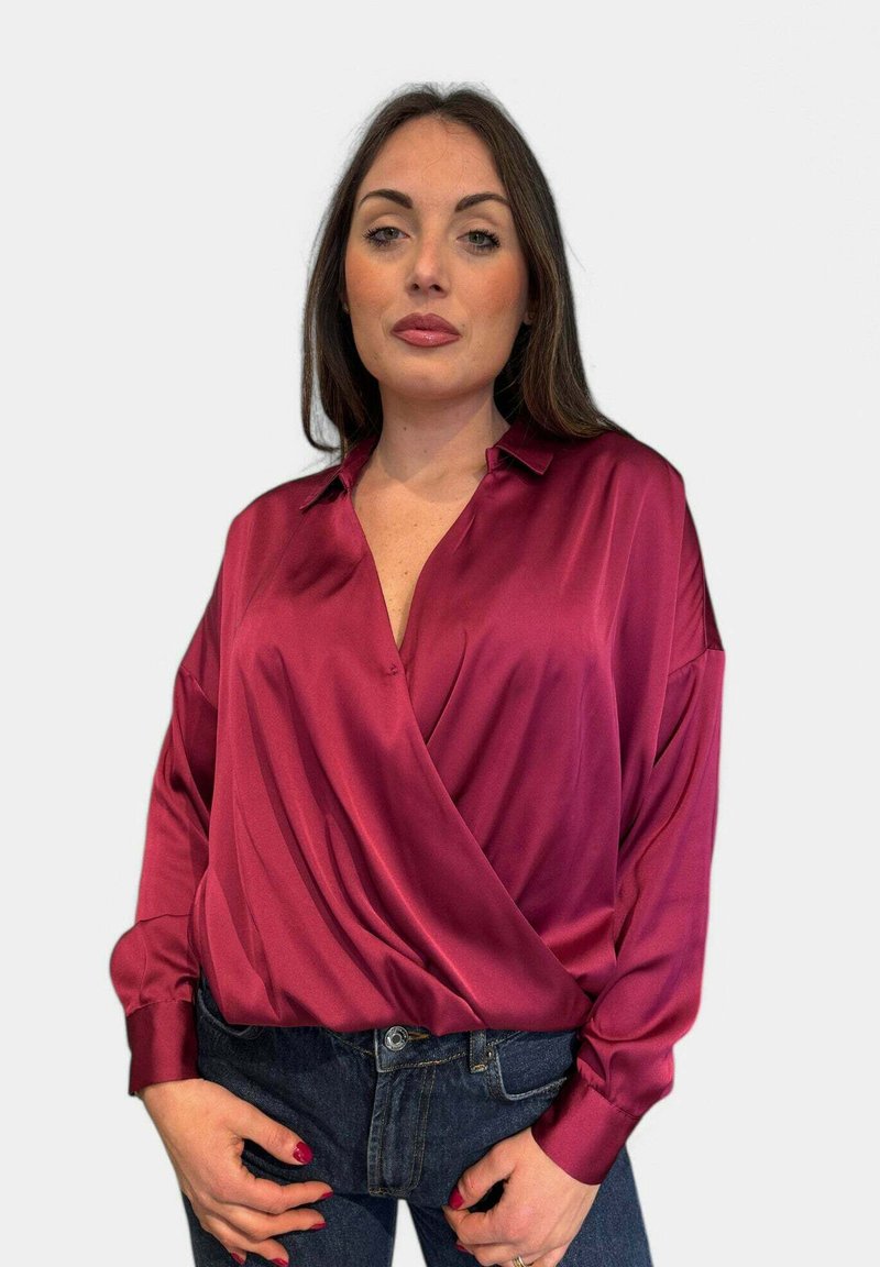 Blusa di raso bordeaux con scollatura a V, maniche lunghe e parte anteriore drappeggiata. Abbinata a jeans blu scuro, che mostrano una superficie liscia e una lucentezza.