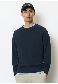 Marineblauer Pullover mit geripptem Muster, Rundhalsausschnitt und langen Ärmeln. Tragen über einem hellblauen Hemd, kombiniert mit einer dunklen Mütze mit weißem Text.
