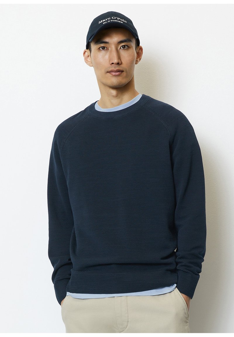 Marineblauwe trui met ribbeltextuur, ronde hals en lange mouwen. Draagt over een lichtblauw shirt, in combinatie met een donkere pet met witte tekst.