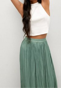 Vrouw met lang donker haar draagt een mouwloos wit crop top en een hoog-getailleerde geplooide saliegroene rok, met haar armen boven haar hoofd geheven.