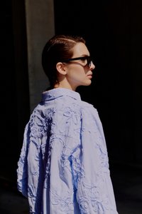 Γυναίκα με κοντά σκούρα μαλλιά που φοράει μαύρα γυαλιά ηλίου και ανοιχτό μπλε μπλούζα με ανάγλυφη κεντημένη λουλουδένια διακόσμηση, κοιτάζοντας δεξιά με σκουρόχρωμο φόντο.