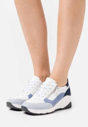 Baskets compensées basses avec panneaux blancs, bleus et gris, lacets à l'avant, fermetures éclair latérales, portées pieds nus sur un fond uni.