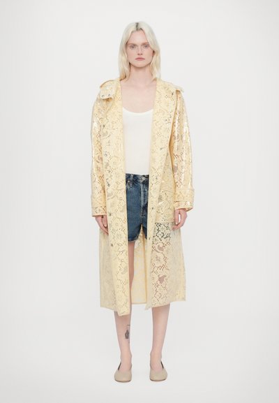 Manteau trench en dentelle jaune avec un design léger et transparent, manches longues et coupe décontractée, porté sur un haut blanc et un short en jean.
