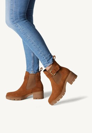 Stivali ankle in camoscio marrone con pannelli laterali elasticizzati, dettaglio fibbia e suola in gomma decorata, indossati con jeans skinny blu.