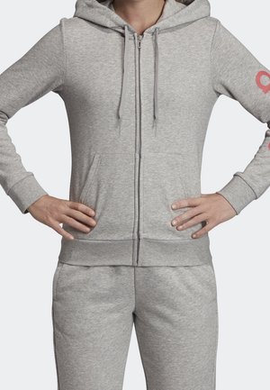 Osoba nosící světle šedou mikinu na zip s kapsami a odpovídající tepláky, ruce opřené o boky, viditelná tvář chybí.