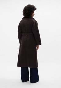 Abrigo de lana marrón y peludo con un amplio cuello, cintura ceñida y mangas largas, combinado con unos jeans acampanados de color azul oscuro. Vista trasera contra un fondo sencillo.