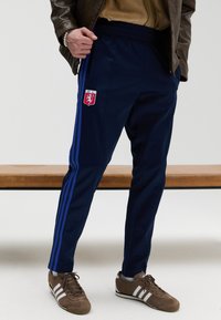 Marineblå treningbukser med elastisk midjebånd, blå side-striper og en logo-patch. Brukt sammen med brune joggesko med hvite striper.